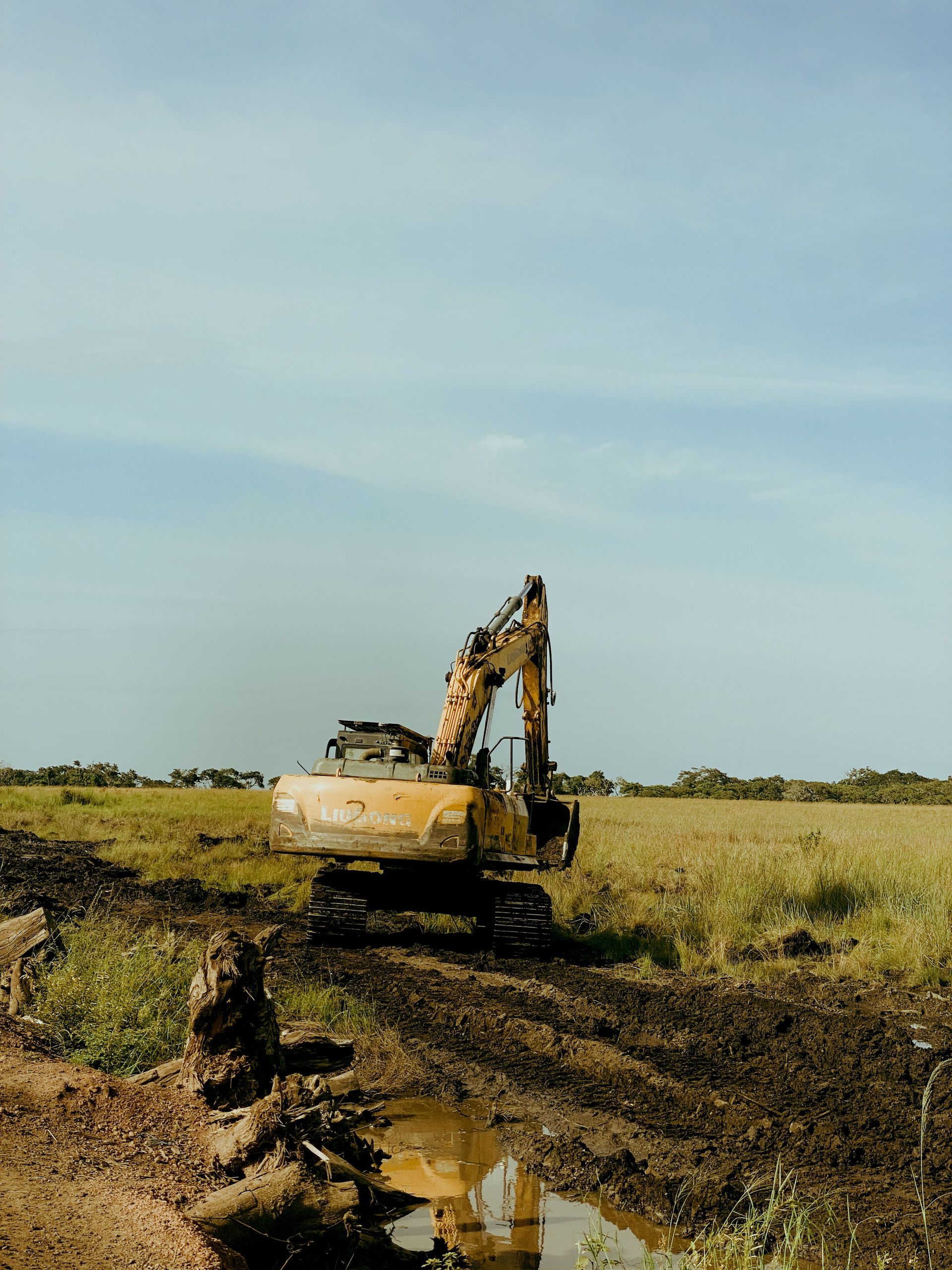 An excavator at work on a rural construction site, surrounded by field and clear sky.